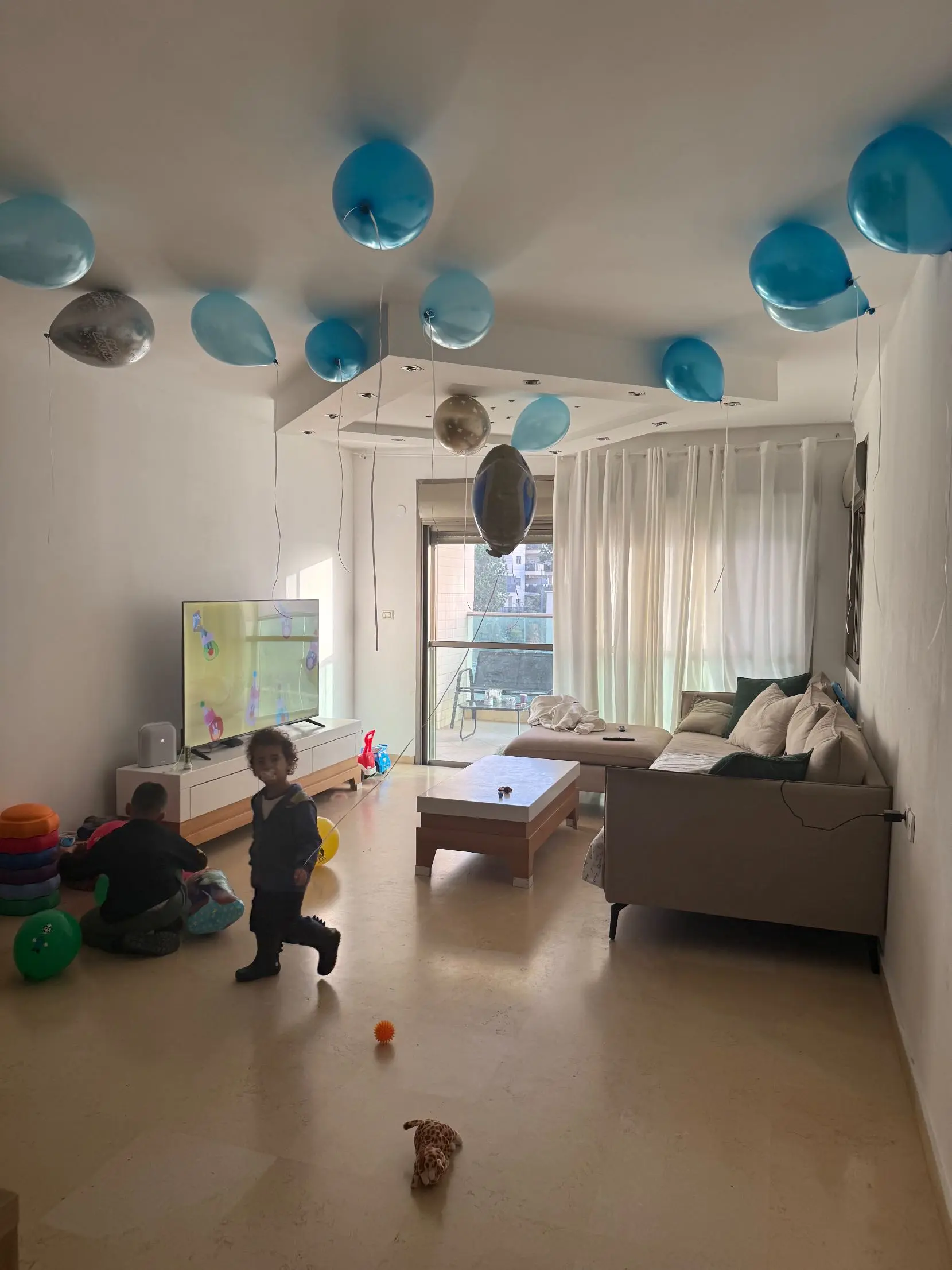 <p>Generater a hyper-realistic image of a bright and airy modern living room embodying Scandinavian minimalism. Dimensions are approximately 5m x 7m. Large, sheer white curtains covering balcony doors illuminate the room, while the walls are painted a warm, soft white. In the center, a large sectional sofa in light gray fabric is decorated with textured cushions in shades of white and pale green. There's a sleek, low-profile media console in light oak wood under a large, modern OLED TV. Above, floating shelves showcase minimalist decor and potted plants. The coffee table is round and composed of light oak, it replaces the previous table. Positioned on top of a soft, plush, neutral-toned area rug that frames the seating area. A dedicated children’s play area is set near the TV comprising of a vibrant, padded play mat, open-shelved toy storage unit, with a petite table accompanied by matching chairs. The room is devoid of balloons. The floor is of light-colored hardwood, aimed to promote a clean, uncluttered, inviting ambiance that facilitates relaxation and family interaction.</p>